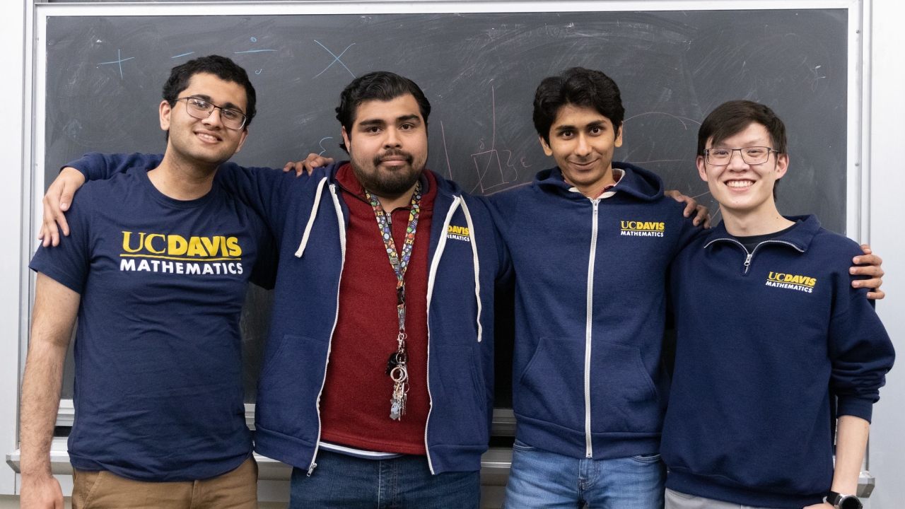 Four members from Math Club at UC Davis pose in front of a black chalkboard. Some members are dressed in T Shirts that have the Math Club logo on them.