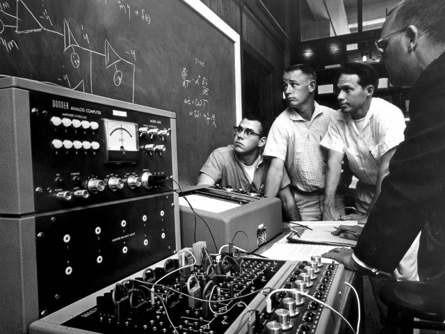 Black and white photo of four UC Davis engineers next to electrical equipment and a chalkboard