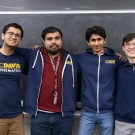 Four members from Math Club at UC Davis pose in front of a black chalkboard. Some members are dressed in T Shirts that have the Math Club logo on them.