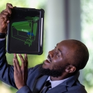 Ismael Mayanja wearing a suit holds up a tablet with a green model displayed